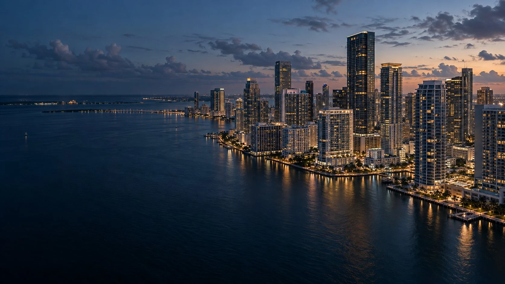 Twilight cityscape showing illuminated skyscrapers along a waterfront at dusk with purple sky and water reflections